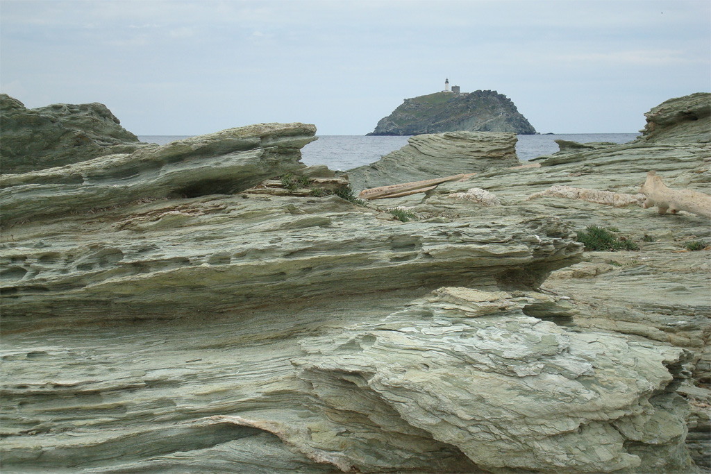 Falaises de prasinites et serpentinites : roches métamorphiques dérivant du gabbro à débit schisteux, riches en amphiboles, épidote, chlorite (schistes verts). Au loin l'îlot de la Giraglia formé lui aussi de prasinite est bien visible.