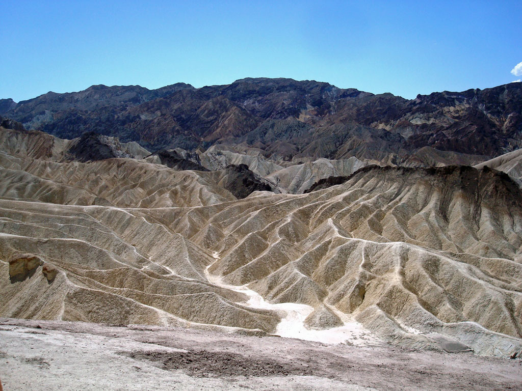 Death Valley (endroit le plus chaud de la planète). La vallée de la mort est un long bassin de 68 km de long et de 8 km de large.
L'histoire géologique des collines et montagnes de Zabriskie Point est impressionnante. Les plus vieilles roches datent de 1,7 milliard d'années, on les trouve dans les Black Mountains à l'arrière plan de la photographie. Au cours de cette longue histoire géologique, la mer s'est retirée, les plaques se sont soulevées, écartées et les failles et l'érosion ont façonné ces magnifiques paysages. Le fond du bassin est recouvert de 3 km de sédiments et de cristaux de sel, le borate (tétraborate de potassium) est une substance naturelle produite par l'évaporation répétée des lacs saisonniers, il est utilisé dans l'industrie des savons et lessives. 