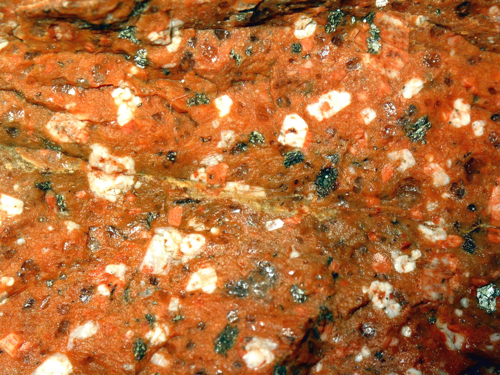 La rhyolite est une roche volcanique rouge ou grise issue d'une lave visqueuse conférant au volcan un caractère explosif.   La macrophotographie (échelle x2) montre distinctement des macro-cristaux isolés de quartz (gris transparents, luisants), de feldspath blanc et de mica noir (biotite). Ces cristaux sont noyés dans une pâte rouge d'apparence non cristallisée. Au microscope, cette pâte montrerait de nombreux microcristaux en forme de baguette, les microlithes.