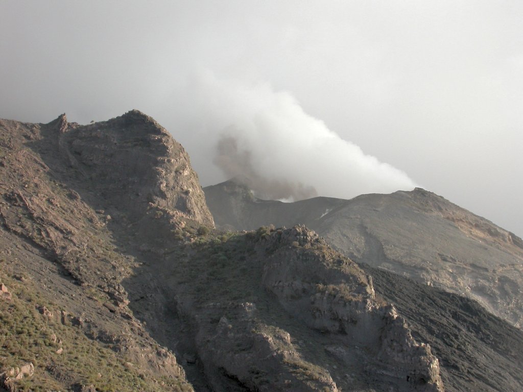 Cratère du Stromboli, dans les îles Eoliennes. Ce volcan présente une intense activité depuis plus de 3 000 ans. Le magma basaltique est très chaud, fluide et assez pauvre en gaz. Lorsque le magma remonte, des bulles se forment par dépressurisation. Les bulles de gaz se séparent spontanément du magma, remontent vers la surface et forment de véritables poches de gaz à quelques mètres sous la surface. Au bout d'un moment, la pression gazeuse devient suffisante pour expulser la colonne de magma qui se trouve au-dessus dans le conduit : c'est ce mécanisme qui est à l'origine des explosions de type strombolien. Les poches de gaz étant séparées les unes des autres, les explosions sont elles-mêmes séparées par des périodes de repos de plusieurs minutes. Le magma éjecté se fragmente en retombant autour du cratère sous forme de projections de taille variable (cendres, lapilli, blocs) qui en s'accumulant construisent les cônes de projections caractéristiques du dynamisme strombolien.
<BR>
<A HREF='https://phototheque.enseigne.ac-lyon.fr/photossql/GoogleEarth/stromboli.kmz'>
<IMG SRC='googleearth.gif' BORDER=0>
</A>