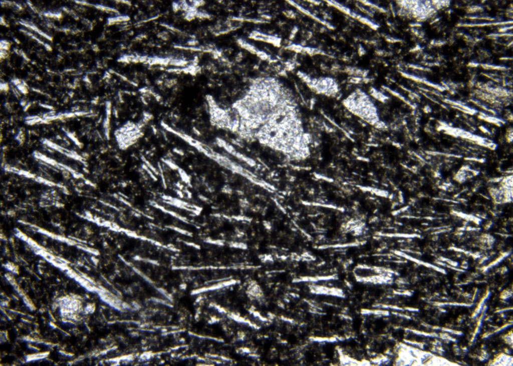 La photographie réalisée au microscope (G=200 x) montre la « pâte » gris bleutée du trachyte. Celle-ci comporte de nombreux microcristaux de feldspath, en forme de baguette. Ces cristaux semblent orientés ce qui peut signifier un écoulement de la lave figé lors du refroidissement. Entre ces microlithes, la matière noire est non cristallisée.